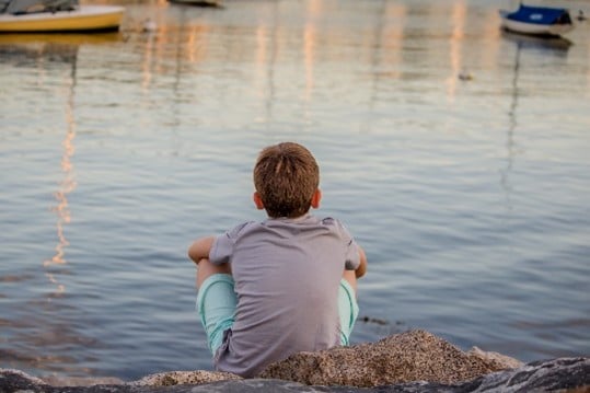 A child sitting by a calm lake, facing away from the camera