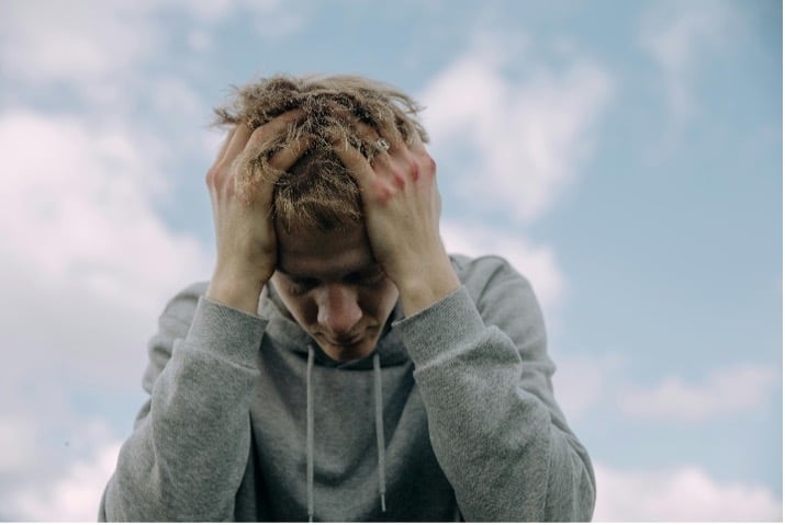 A person wearing a grey sweater standing outdoors with their head down, holding their head in their hands